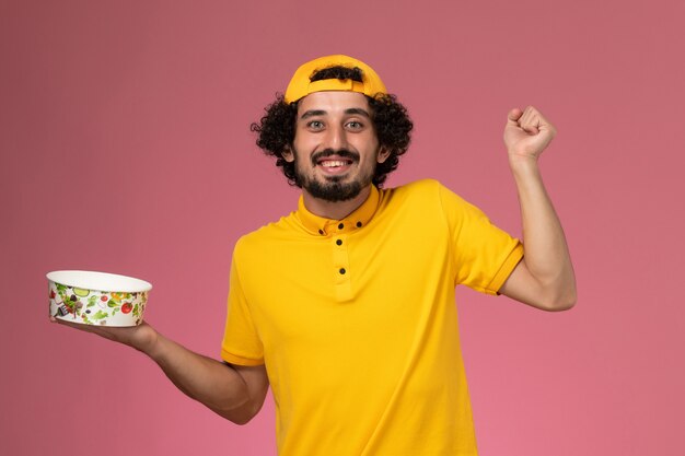 Corriere maschio di vista frontale in uniforme gialla e mantello con ciotola di consegna rotonda sulle sue mani che si rallegrano sullo sfondo rosa.