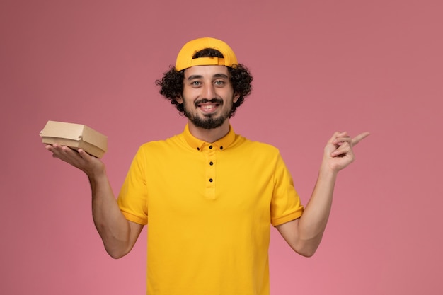 Corriere maschio di vista frontale in uniforme gialla e mantello con piccolo pacchetto di cibo di consegna sulle mani sullo sfondo rosa.