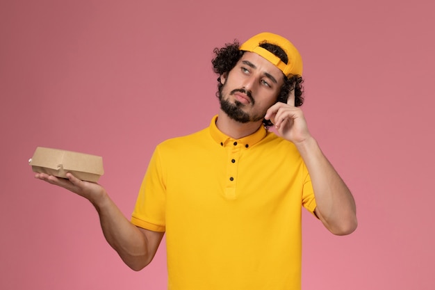 Corriere maschio di vista frontale in uniforme gialla e mantello con poco pacchetto di cibo di consegna sulle sue mani pensando sullo sfondo rosa.