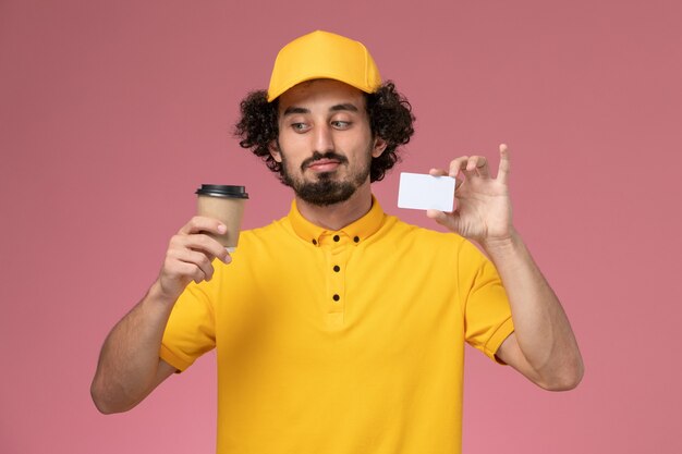 Corriere maschio di vista frontale in uniforme gialla e tazza di caffè di consegna della tenuta del capo e carta sulla parete rosa