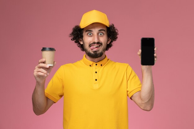 Corriere maschio di vista frontale in uniforme gialla e tazza di caffè di consegna della tenuta del capo e telefono sulla parete rosa