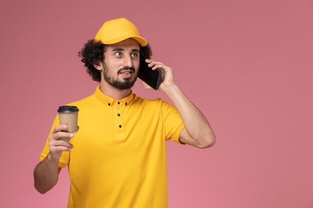 Corriere maschio di vista frontale in uniforme gialla e tazza di caffè di consegna della tenuta del capo e telefono sulla parete rosa