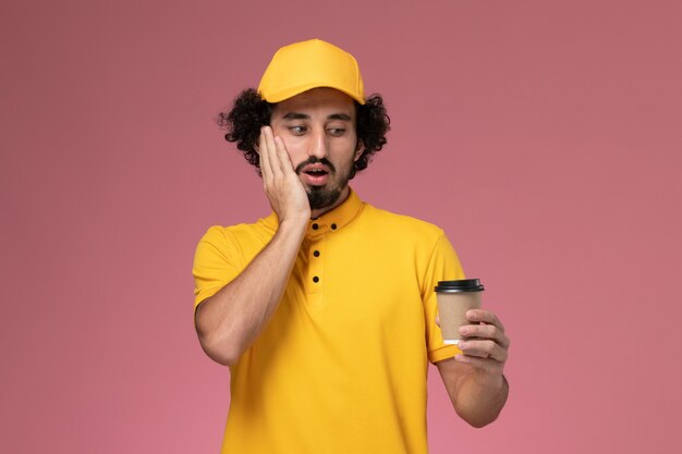 Corriere maschio di vista frontale in uniforme gialla e tazza di caffè di consegna della tenuta del capo sul lavoro dell'azienda di servizio di consegna dell'uniforme dello scrittorio rosa