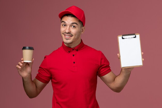 Corriere maschio di vista frontale in uniforme rossa che tiene la tazza di caffè di consegna e blocco note sul lavoro maschio dell'operaio rosa della parete