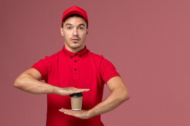 Corriere maschio di vista frontale in uniforme rossa che tiene la tazza di caffè marrone di consegna sul lavoro uniforme maschio dell'operaio di consegna di servizio della parete rosa