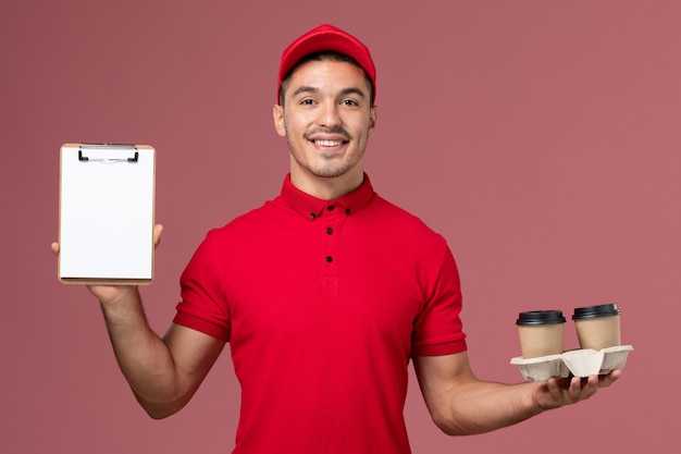 Corriere maschio di vista frontale in uniforme rossa che tiene le tazze di caffè e il blocchetto per appunti marroni di consegna sul lavoratore della parete rosa-chiaro