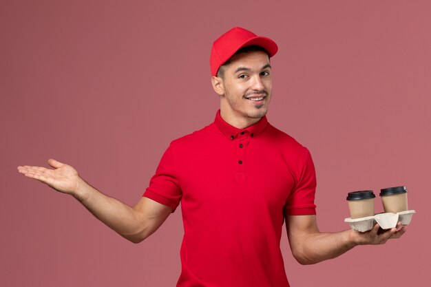 Corriere maschio di vista frontale in uniforme rossa che tiene le tazze di caffè marroni di consegna che sorridono sul lavoratore maschio dello scrittorio rosa