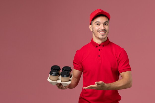 Corriere maschio di vista frontale in uniforme rossa che tiene le tazze di caffè marroni di consegna con il sorriso sul lavoratore rosa della parete