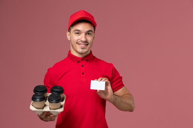 Corriere maschio di vista frontale in uniforme rossa che tiene le tazze di caffè marroni di consegna con la carta bianca sull'uniforme dell'operaio di lavoro di consegna di servizio della parete rosa