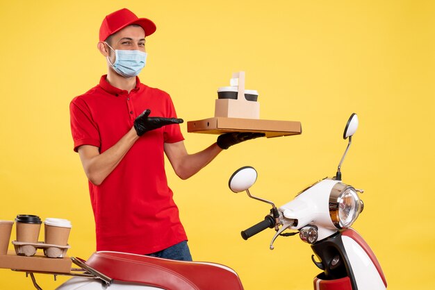 Corriere maschio di vista frontale in uniforme rossa con scatola di cibo e caffè su un virus dell'uniforme di lavoro di covid pandemia di colore di lavoro giallo