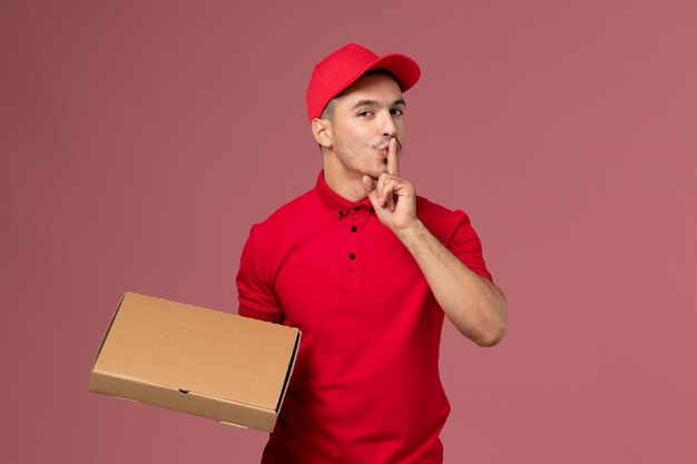 Corriere maschio di vista frontale in uniforme rossa e contenitore di consegna del cibo della tenuta del capo che mostra il segno di silenzio sul lavoro dell'operaio dell'uniforme di consegna rosa dello scrittorio