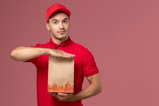 Corriere maschio di vista frontale in uniforme rossa e mantello che tiene il pacchetto di cibo di carta sul muratore rosa