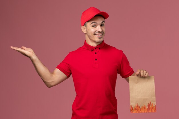 Corriere maschio di vista frontale in uniforme rossa e mantello che tiene il pacchetto di cibo di carta sulla parete rosa