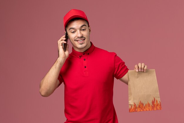 Corriere maschio di vista frontale in uniforme rossa e mantello che tiene il pacchetto di cibo e parla al telefono sulla parete rosa