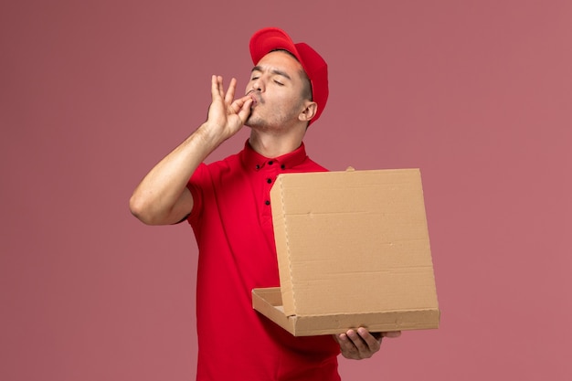 Corriere maschio di vista frontale in uniforme rossa e mantello che tiene la scatola di cibo aprendolo sul lavoro di lavoratore scrivania rosa chiaro