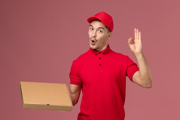 Corriere maschio di vista frontale in uniforme rossa e mantello che tiene la scatola di consegna del cibo sul lavoro di operaio muro rosa chiaro