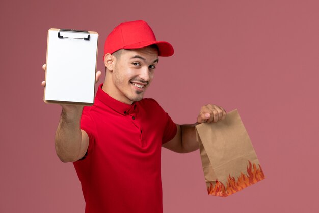 Corriere maschio di vista frontale in uniforme rossa e mantello che tiene pacchetto alimentare e blocco note sorridente sulla parete rosa