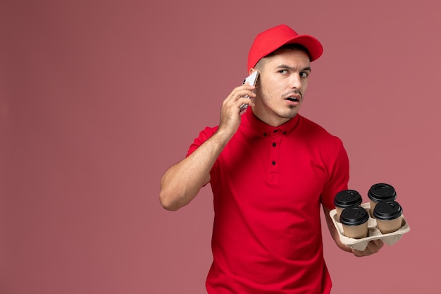 Corriere maschio di vista frontale in uniforme rossa e tazze di caffè di consegna della tenuta del capo che comunicano sul telefono sulla parete rosa