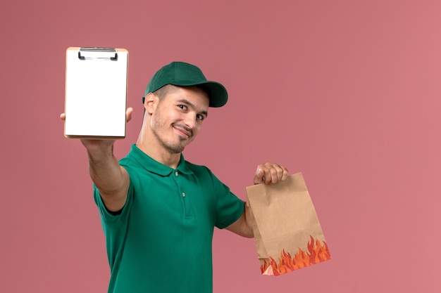 Corriere maschio di vista frontale in uniforme verde che tiene il pacchetto alimentare e il blocco note con il sorriso su sfondo rosa chiaro