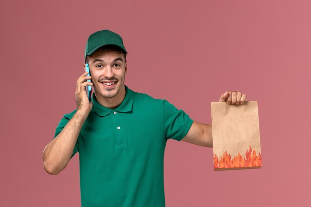 Corriere maschio di vista frontale in uniforme verde che tiene il pacchetto alimentare mentre si parla al telefono in rosa