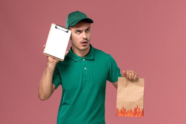 Corriere maschio di vista frontale in uniforme verde che tiene il pacchetto di cibo e il blocco note pensando su sfondo rosa chiaro