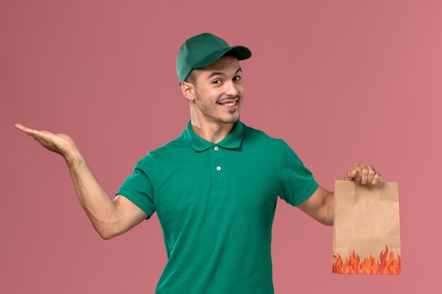 Corriere maschio di vista frontale in uniforme verde che tiene il pacchetto di cibo e sorridente sullo sfondo rosa chiaro