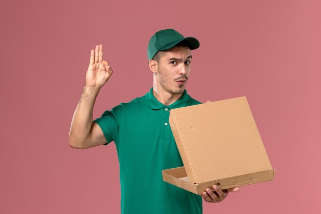 Corriere maschio di vista frontale in uniforme verde che tiene la scatola di cibo e aprendolo su sfondo rosa chiaro