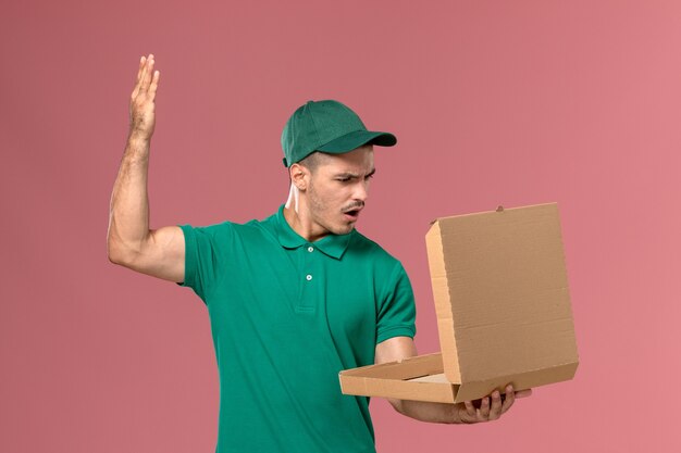 Corriere maschio di vista frontale in uniforme verde che tiene la scatola di cibo e aprendolo su sfondo rosa uniforme di consegna del lavoro maschile di servizio
