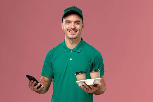 Corriere maschio di vista frontale in uniforme verde che tiene le tazze di caffè di consegna marroni e utilizzando un telefono con il sorriso sulla scrivania rosa