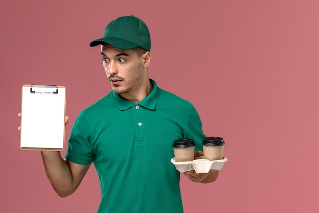 Corriere maschio di vista frontale in uniforme verde che tiene le tazze di caffè e il blocco note marroni di consegna sullo scrittorio rosa-chiaro