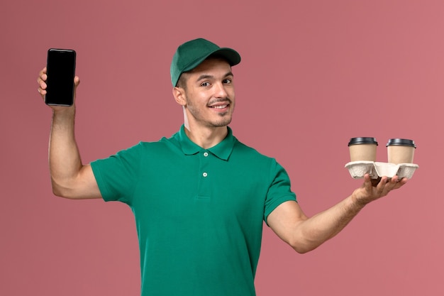 Corriere maschio di vista frontale in uniforme verde che tiene le tazze di caffè e il telefono marroni di consegna con il sorriso sullo scrittorio rosa