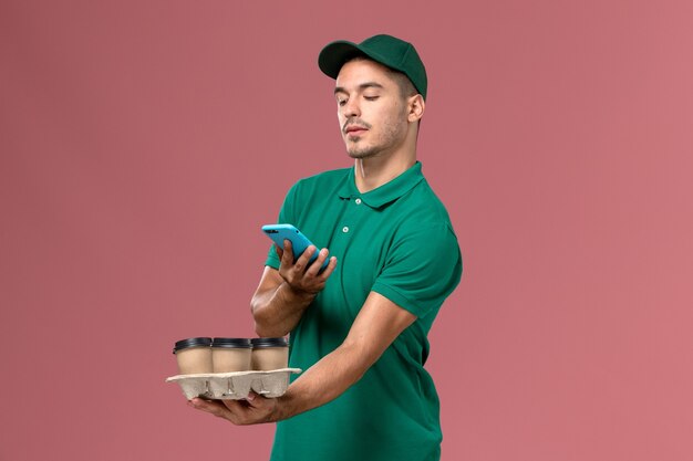 Corriere maschio di vista frontale in uniforme verde che tiene le tazze di caffè marroni che prendono la foto su fondo rosa
