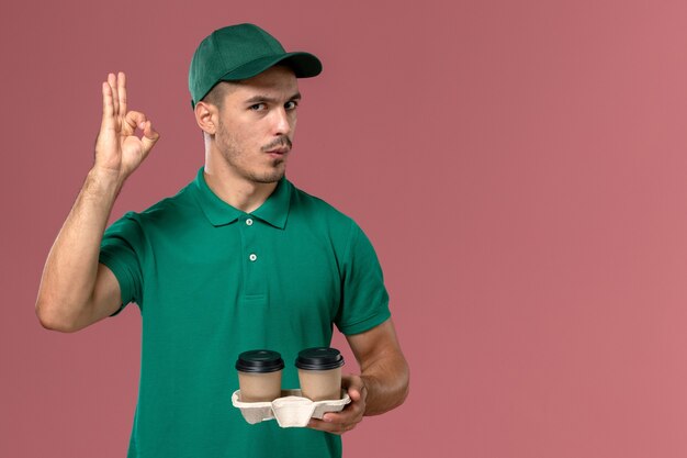 Corriere maschio di vista frontale in uniforme verde che tiene le tazze di caffè marroni di consegna sul maschio rosa dello scrittorio