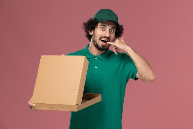 Corriere maschio di vista frontale in uniforme verde e contenitore dell'alimento di consegna della tenuta del capo aprendolo sulla consegna dell'uniforme del lavoratore di servizio del fondo rosa