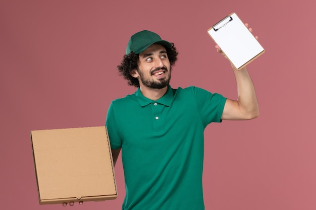 Corriere maschio di vista frontale in uniforme verde e mantello che tiene il blocco note della scatola dell'alimento di consegna sul lavoro di consegna dell'uniforme di servizio del fondo rosa