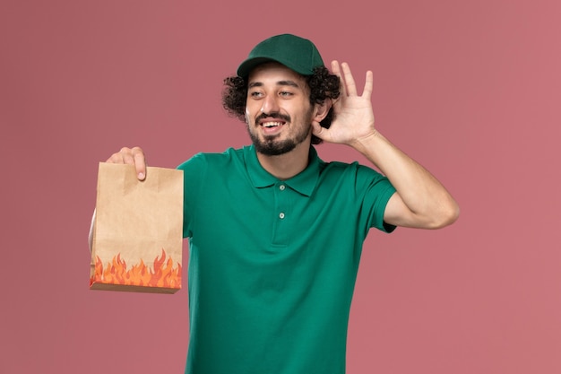 Corriere maschio di vista frontale in uniforme verde e mantello che tiene il pacchetto di cibo di carta cercando di sentire sul lavoro di consegna uniforme del lavoratore di servizio sfondo rosa