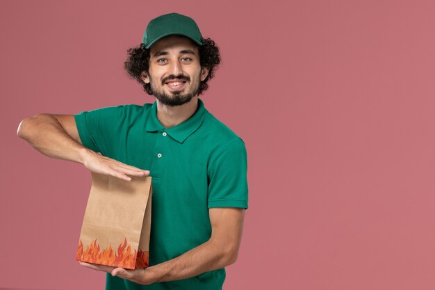 Corriere maschio di vista frontale in uniforme verde e mantello che tiene il pacchetto di cibo di carta con un leggero sorriso sul lavoro di consegna uniforme del lavoratore di servizio di sfondo rosa