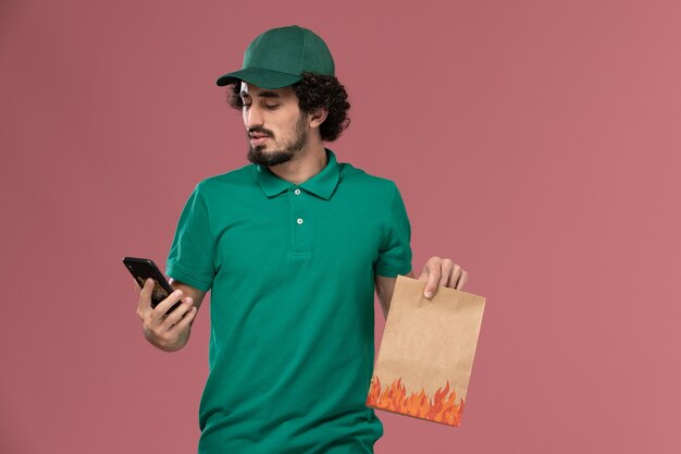 Corriere maschio di vista frontale in uniforme verde e mantello che tiene il pacchetto di cibo e utilizzando il suo telefono sul lavoro maschile di consegna uniforme di servizio sfondo rosa