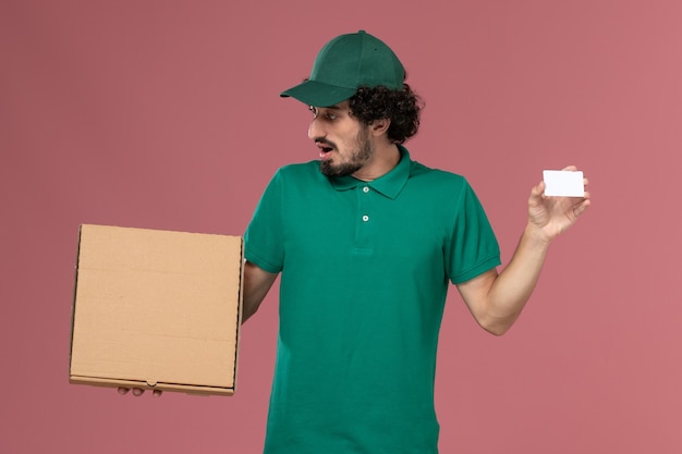 Corriere maschio di vista frontale in uniforme verde e mantello che tiene la scatola di cibo di consegna e la carta sul lavoro di lavoro di consegna uniforme di servizio del pavimento rosa