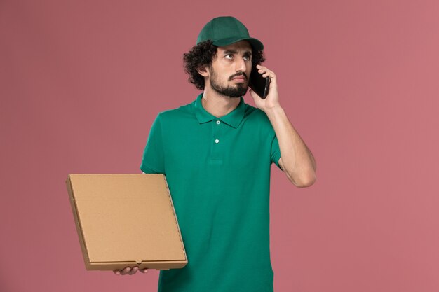Corriere maschio di vista frontale in uniforme verde e mantello che tiene la scatola di cibo per la consegna e parla al telefono sul lavoro di consegna uniforme del lavoratore di servizio del pavimento rosa