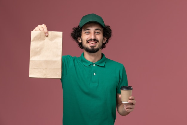 Corriere maschio di vista frontale in uniforme verde e mantello che tiene la tazza di caffè di consegna e il pacchetto di cibo sul lavoro di servizio di consegna uniforme sfondo rosa