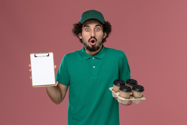 Corriere maschio di vista frontale in uniforme verde e mantello che tiene le tazze di caffè con il blocco note sul maschio dell'operaio di consegna dell'uniforme di servizio del fondo rosa