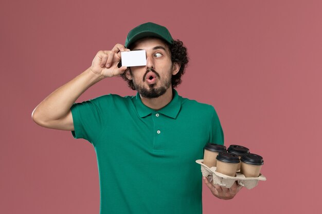 Corriere maschio di vista frontale in uniforme verde e mantello che tiene le tazze di caffè con la carta sul maschio di lavoro dell'operaio di consegna dell'uniforme di servizio del fondo rosa