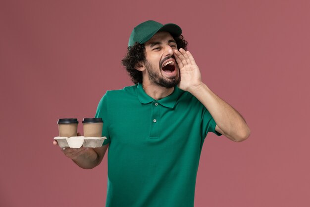 Corriere maschio di vista frontale in uniforme verde e mantello che tiene le tazze di caffè di consegna che grida sulla consegna dell'uniforme maschio di lavoro di servizio del fondo rosa