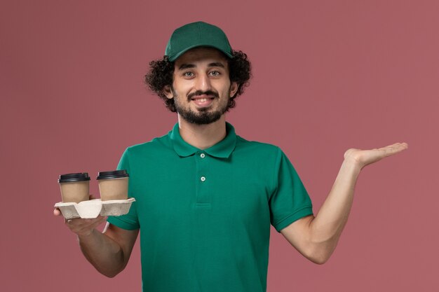 Corriere maschio di vista frontale in uniforme verde e mantello che tiene le tazze di caffè di consegna con il sorriso sul lavoro di lavoro di consegna dell'uniforme di servizio del fondo rosa