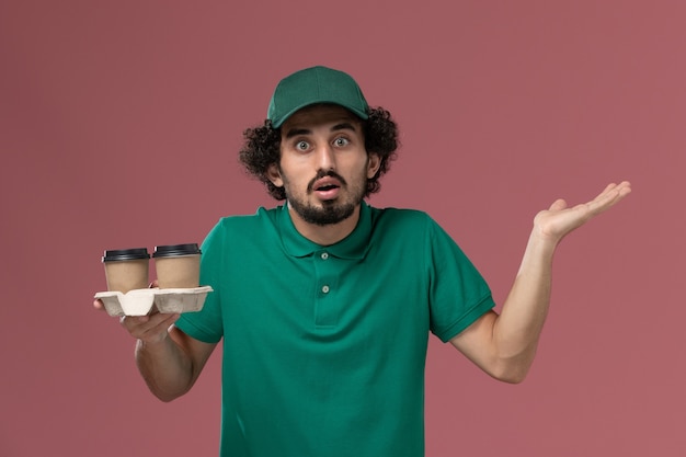 Corriere maschio di vista frontale in uniforme verde e mantello che tiene le tazze di caffè di consegna su fondo rosa servizio di consegna uniforme lavoro lavoratore maschio lavoro