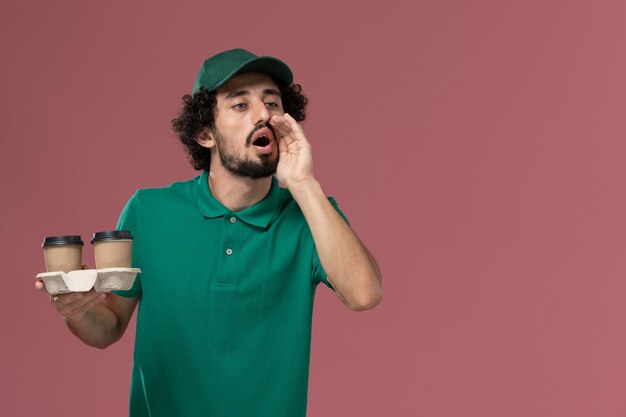 Corriere maschio di vista frontale in uniforme verde e mantello che tiene le tazze di caffè di consegna sulla consegna uniforme maschile di servizio di lavoro di sfondo rosa