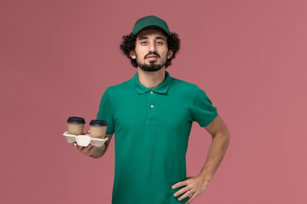 Corriere maschio di vista frontale in uniforme verde e mantello che tiene le tazze di caffè sul lavoro di lavoratore di servizio di consegna uniforme sfondo rosa chiaro