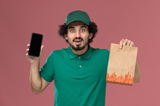 Corriere maschio di vista frontale in uniforme verde e mantello che tiene pacchetto di cibo e smartphone sul lavoro maschile di consegna uniforme di servizio sfondo rosa