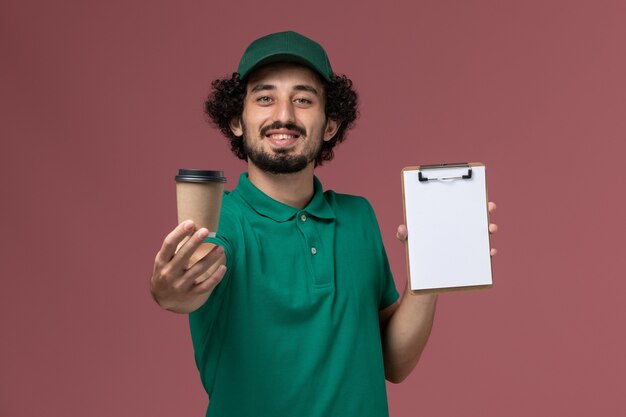 Corriere maschio di vista frontale in uniforme verde e tazza di caffè di consegna della tenuta del capo con il blocco note e il sorriso sul lavoro dell'azienda di servizio di consegna dell'uniforme dello scrittorio rosa